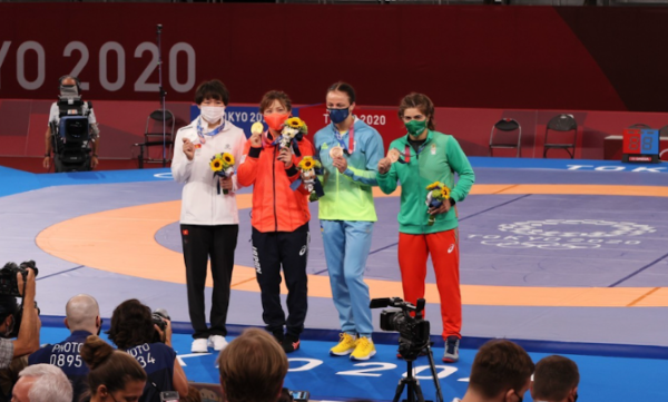 Four athletes stand to greet the media following their freestyle wrestling performances at the 2020 Tokyo Olympics, which were actually played in 2021. They each wear a different color medal depending on how they placed in the contest. 