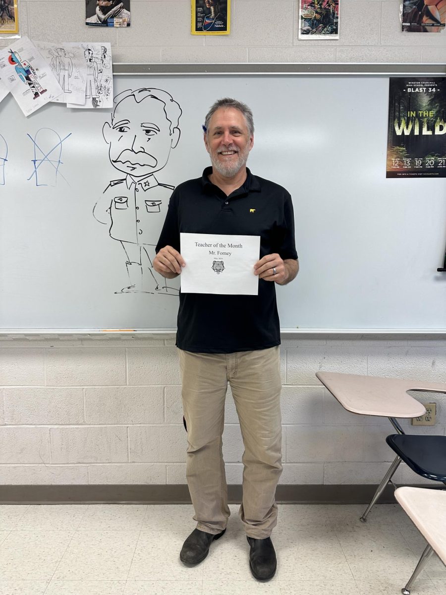 Mr. Christopher Forney smiles while he stands in front of a white board drawing of himself, holding his teacher of the month certificate. He has been teaching MCPS students in subjects such as psychology and world history for 26 years.